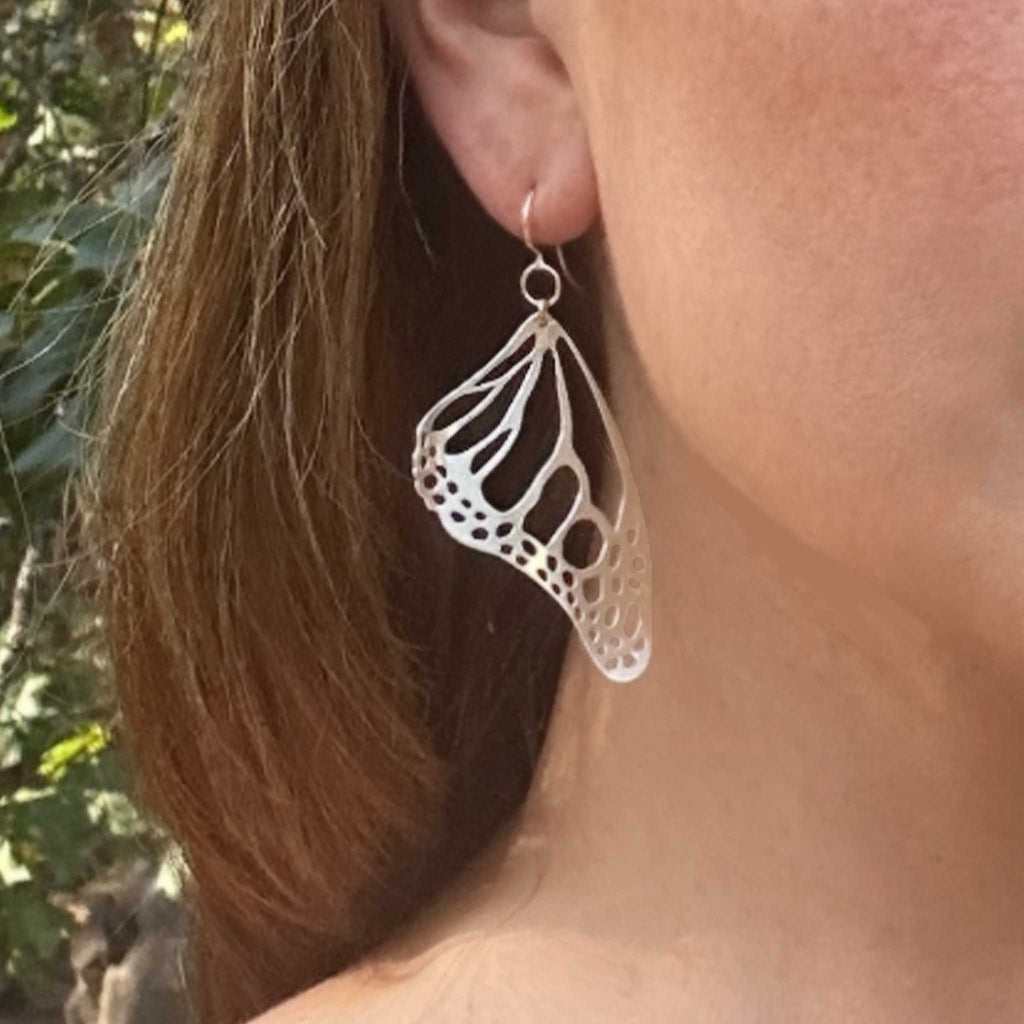 Close-up of a person wearing Monarch silver butterfly-shaped earring with a blurred natural background.