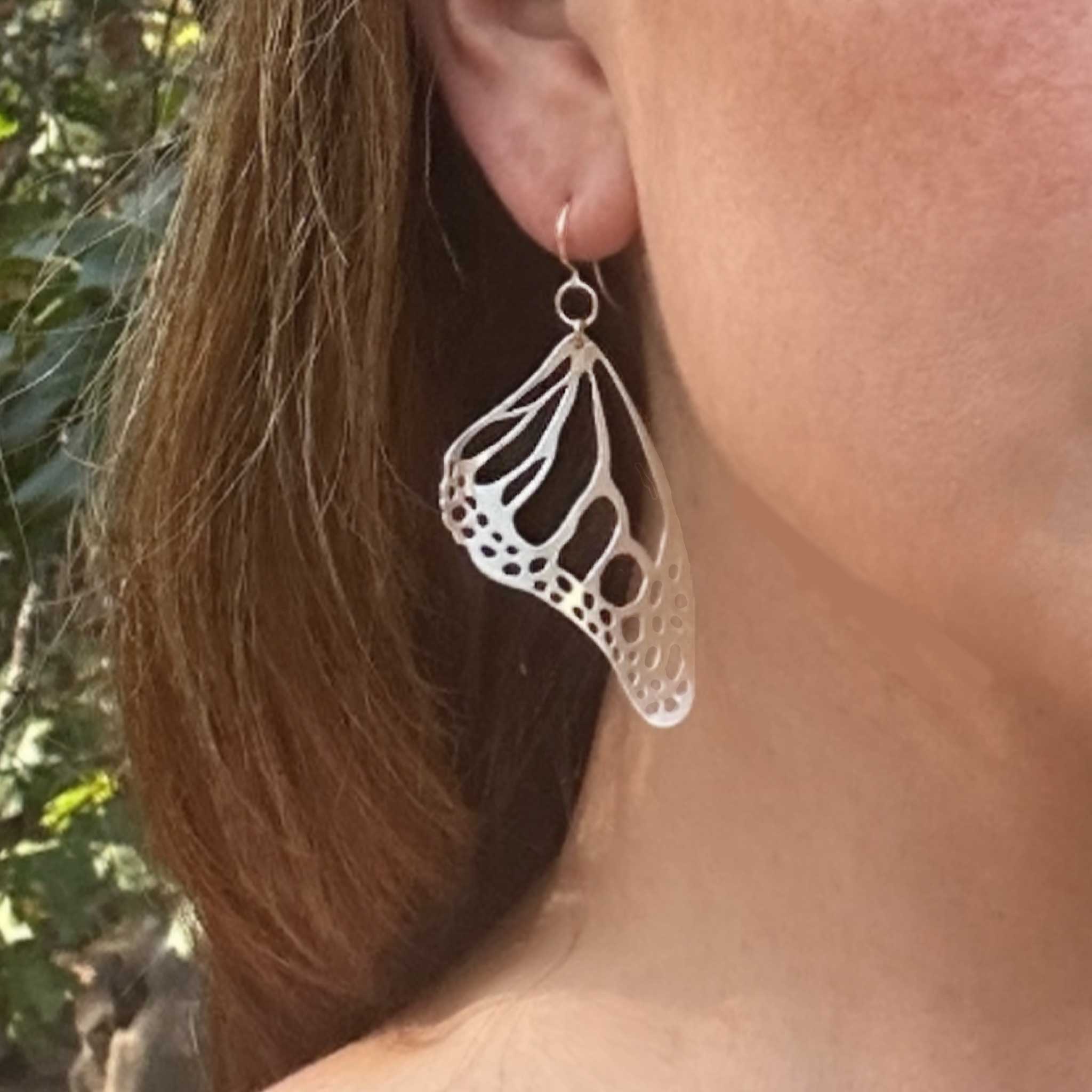 Close-up of a person wearing Monarch silver butterfly-shaped earring with a blurred natural background.