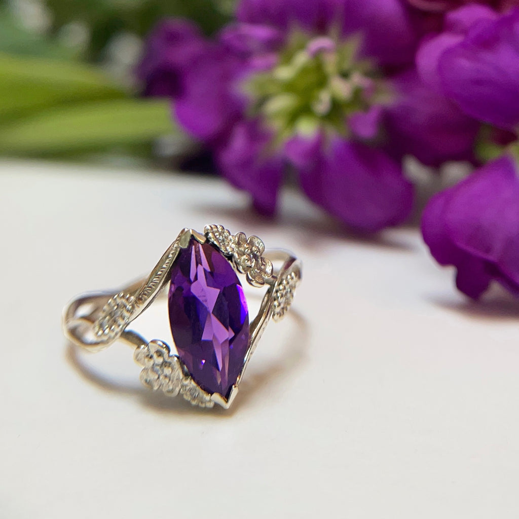 Custom colored stone engagement ring with a purple gem in  floral vines in 18k white gold in front of purple flowers | Camille Torres Designs