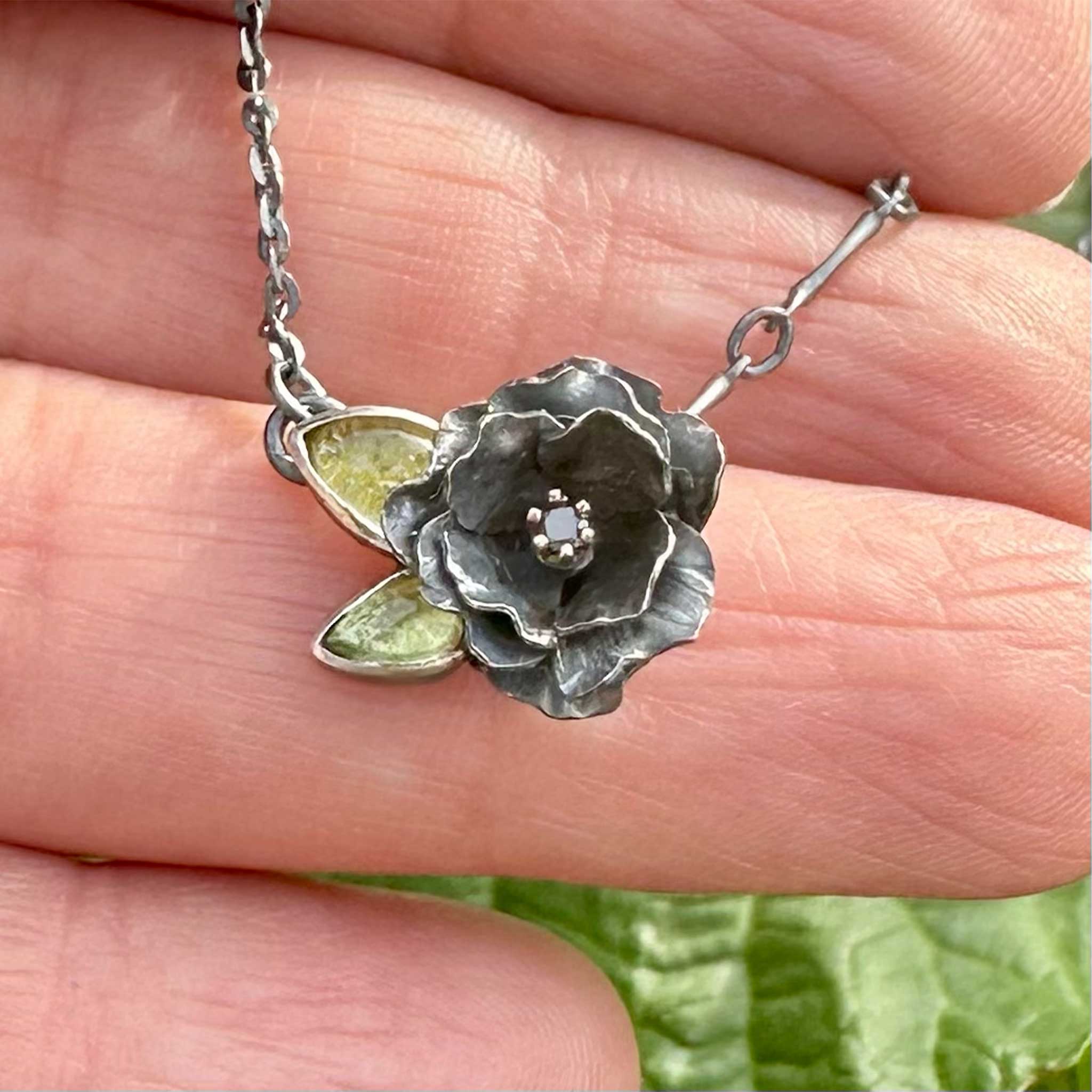 Silver oxidized flower-shaped necklace with green gemstones leaves and a black diamond center set in 14k gold, held between fingers against a blurred green background
