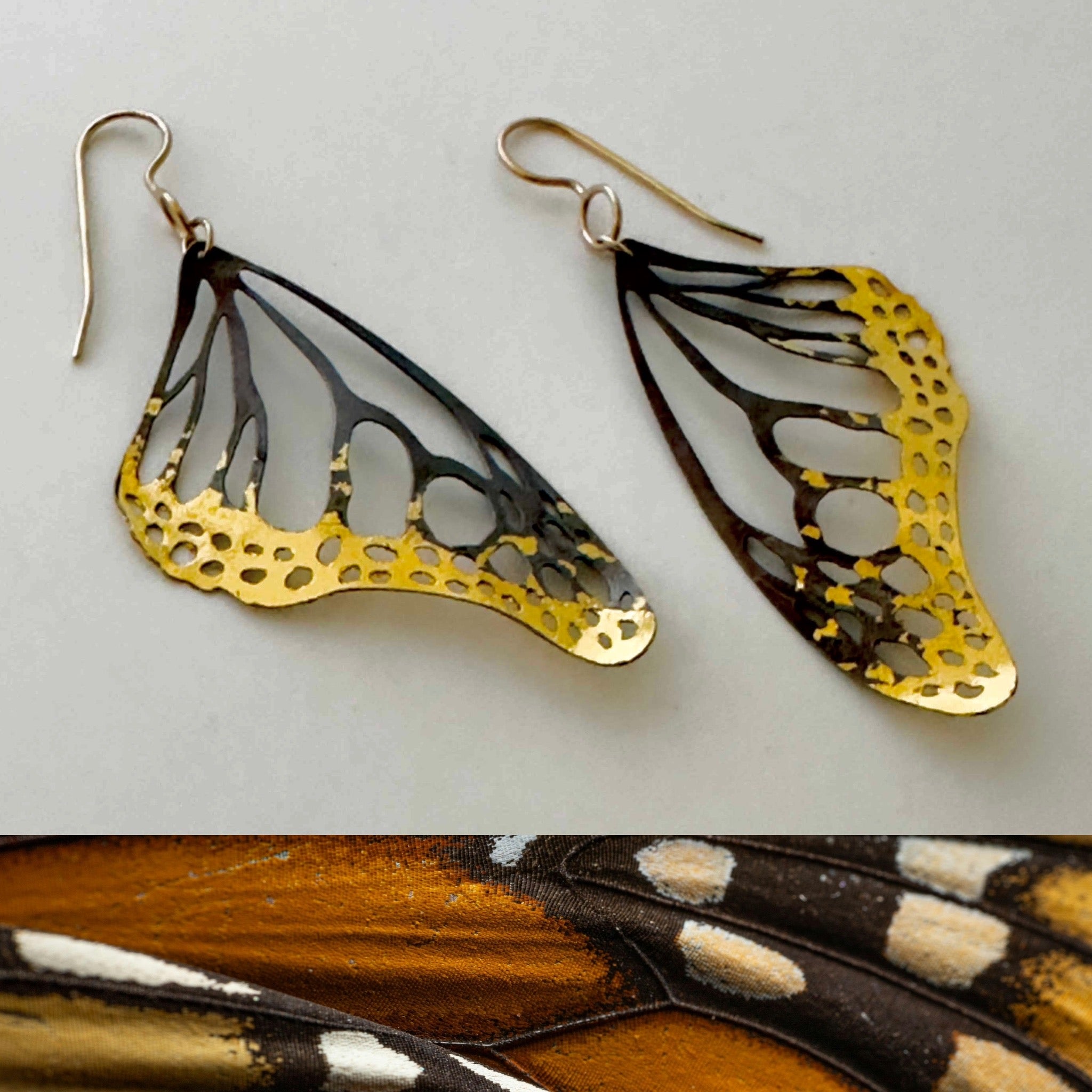 Butterfly wing-shaped earrings with 24k gold and black details on a light gray background.