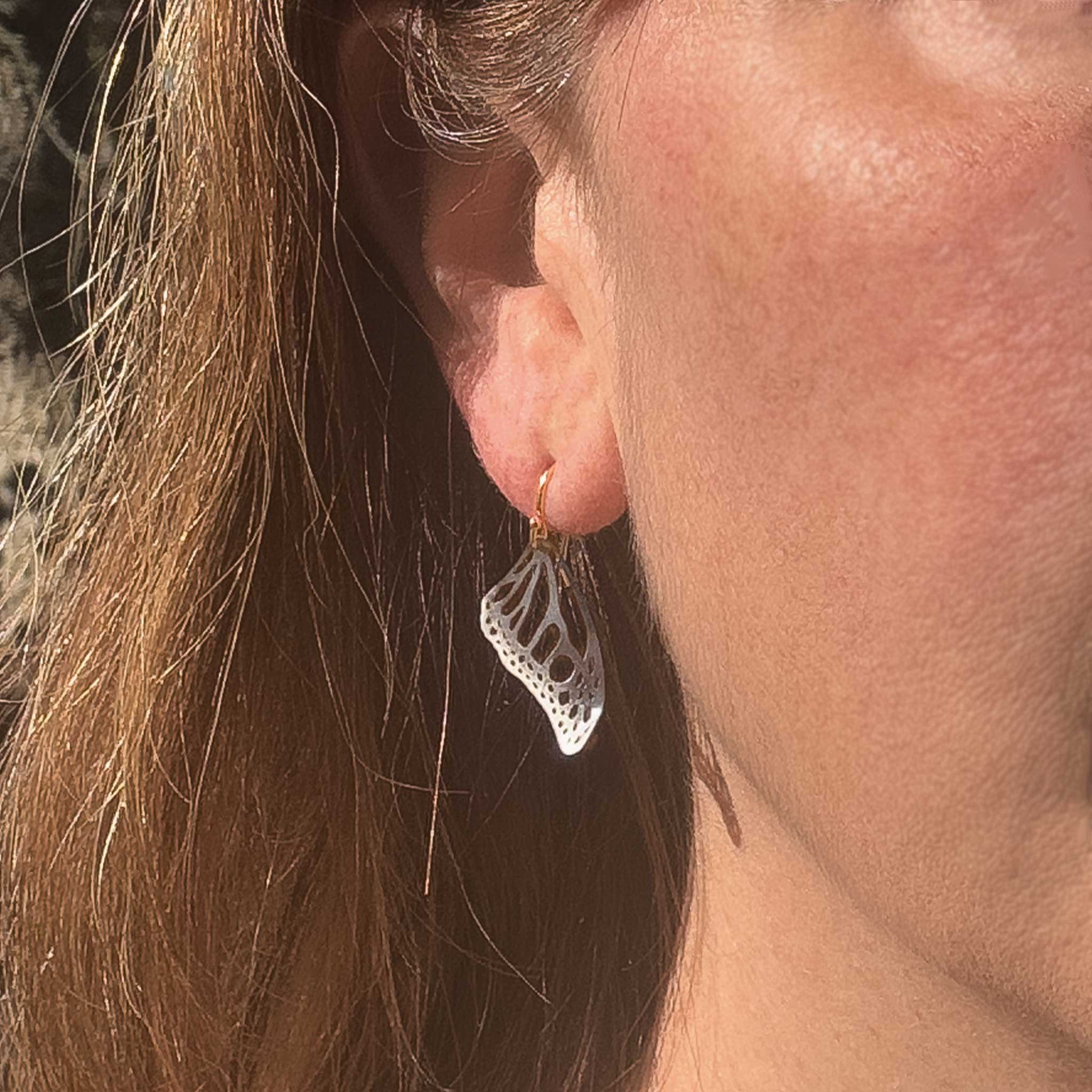 Close up of person wearing small Monarch butterfly drop earrings in oxidized silver with 14k gold ear wires over a white background | Camille Torres Designs