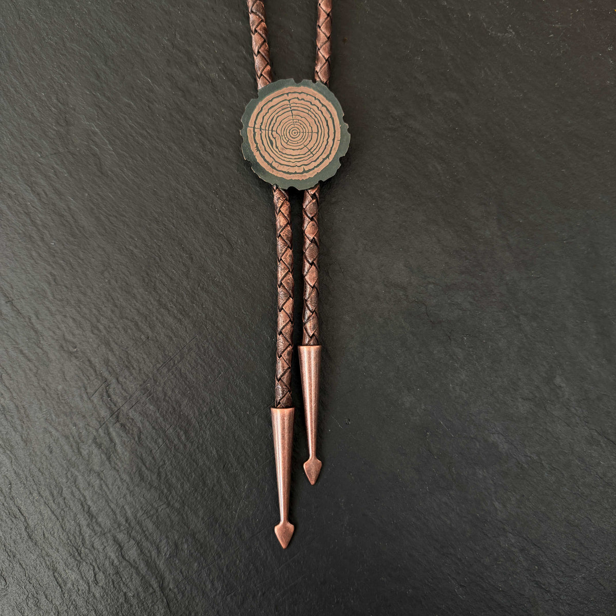 Redwood Slice Bolo Tie, etched copper tree-ring slide on Dark Brown braided leather with copper arrowhead tips on a slate background. | Camille Torres Designs