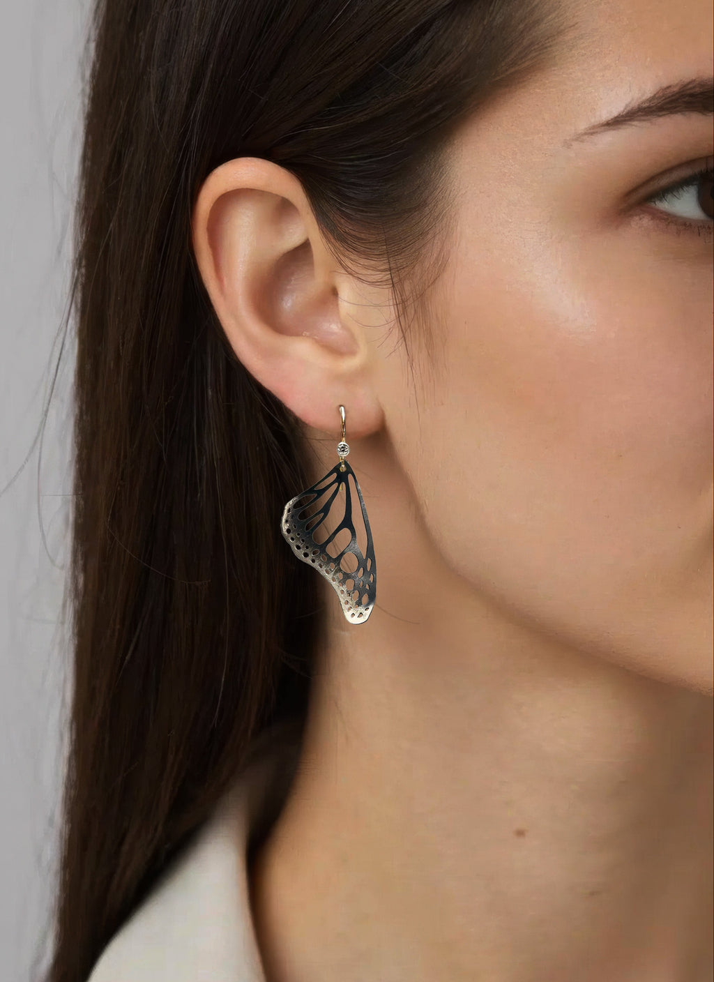 A close-up of a woman wearing a delicate oxidized silver Monarch butterfly dangle earring, accented with a diamond and 14k gold ear wire