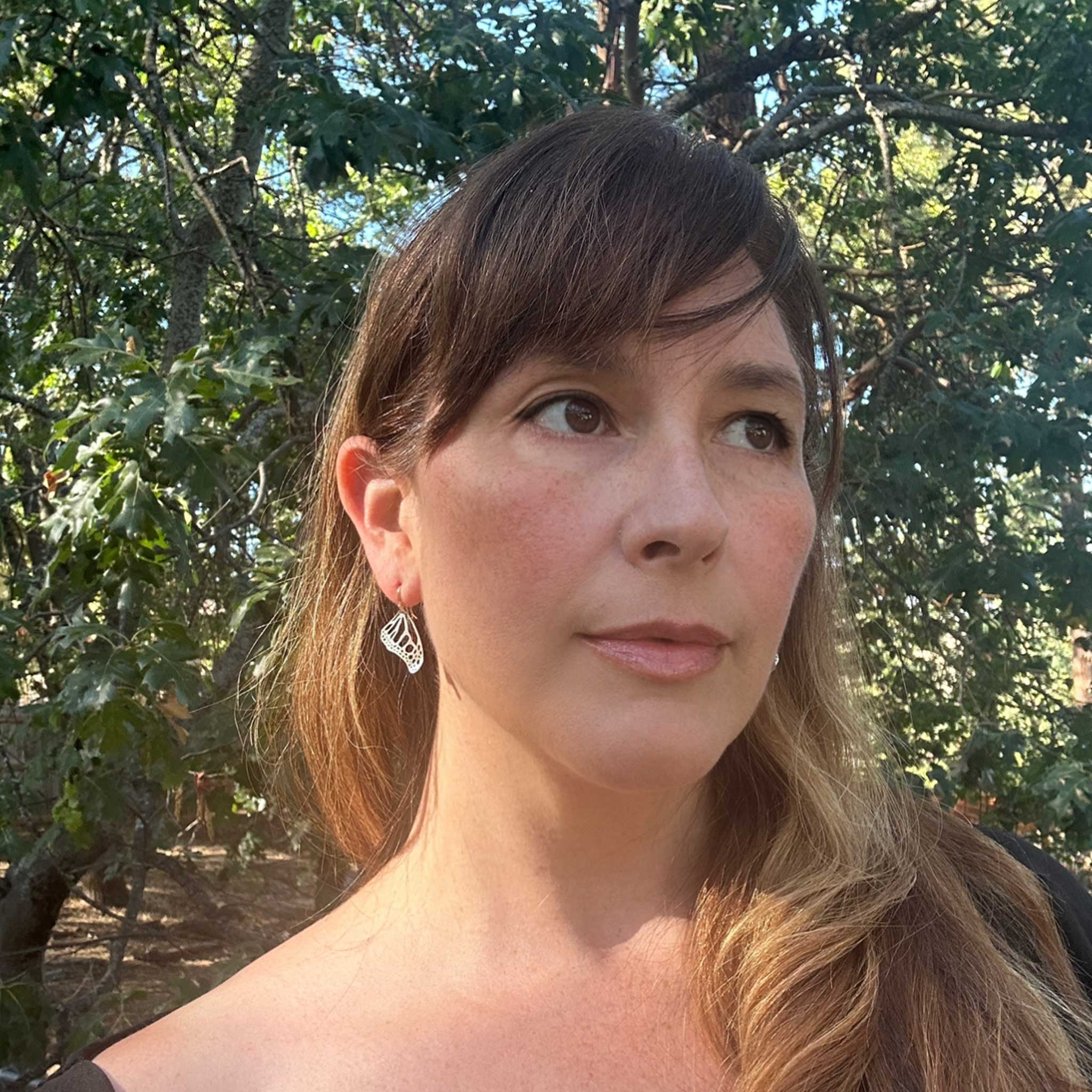 Camille, with long brown hair and small Monarch silver earrings, standing outdoors with trees in the background