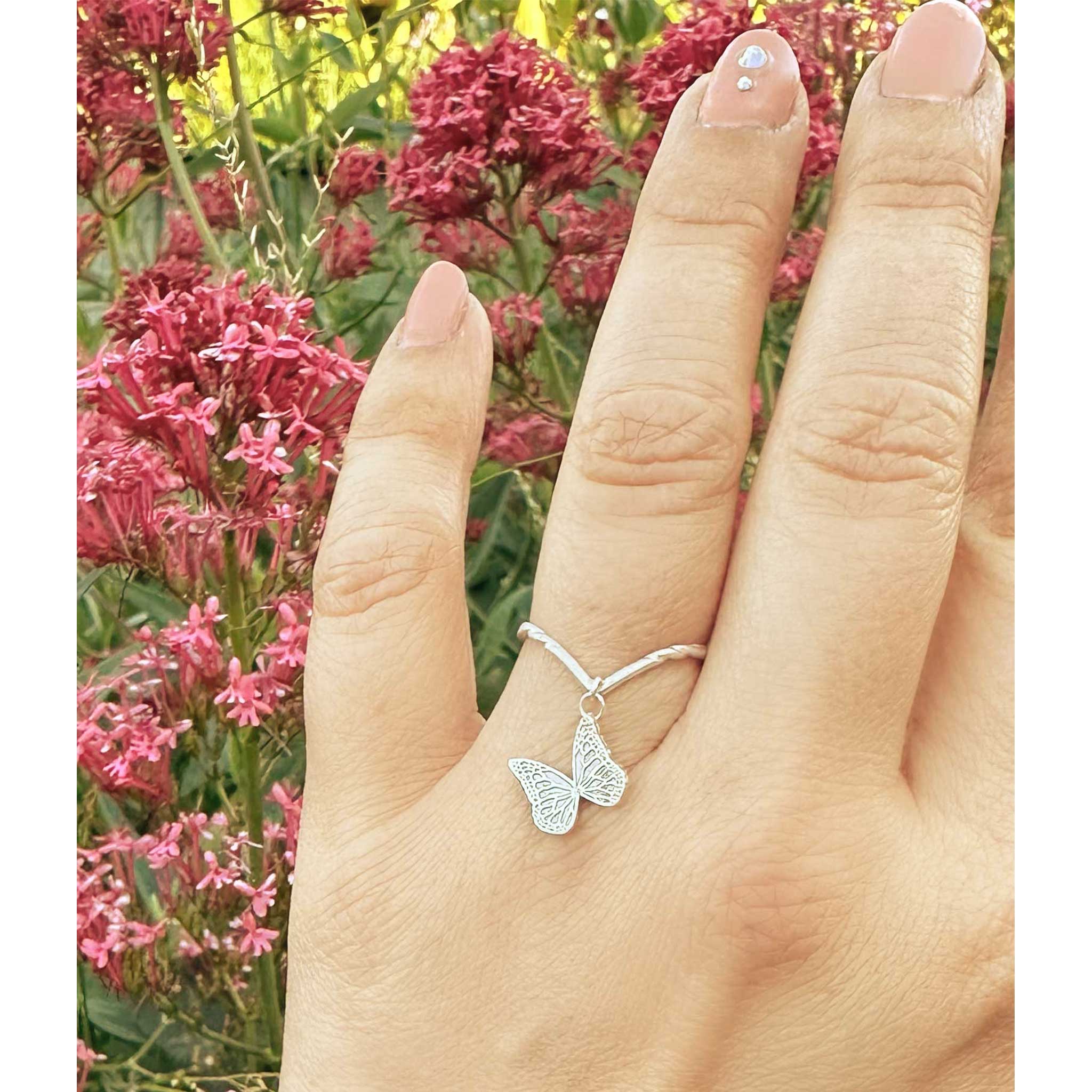 Hand wearing a Whisper silver butterfly charm ring with pink flowers in the background